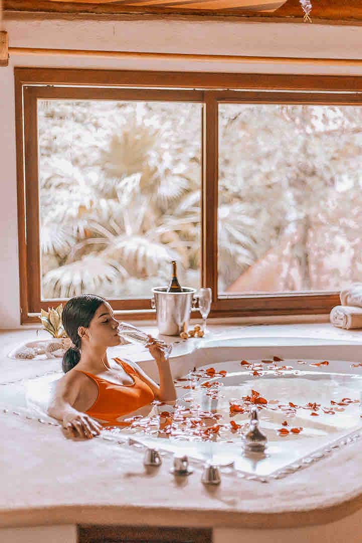 A relaxing jacuzzi is set against a backdrop of large windows, allowing natural light to flood the space. Rose petals are scattered across the water's surface. A bottle of champagne and two glasses are positioned nearby, enhancing the tranquil ambiance.