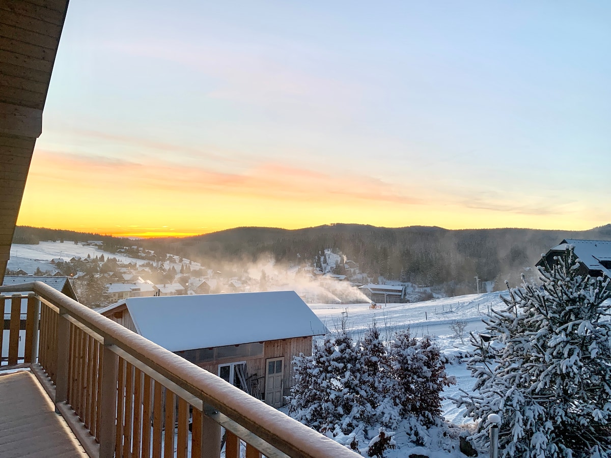 A tranquil winter landscape is captured from a spacious wooden terrace. Snow blankets the ground and nearby trees, while distant hills are illuminated by the soft glow of a sunrise, enhancing the serene atmosphere of the scenic surroundings.