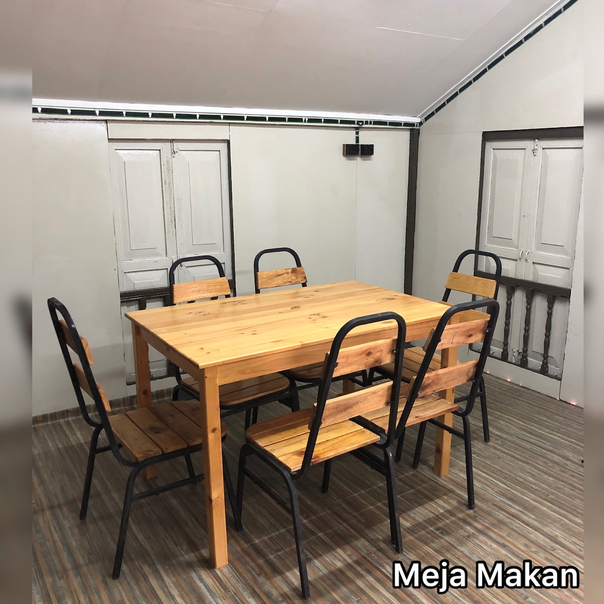 A wooden dining table is surrounded by six black metal chairs with wooden seating. The room features two closed white doors and a simple wall design, creating a functional dining space suitable for group gatherings.