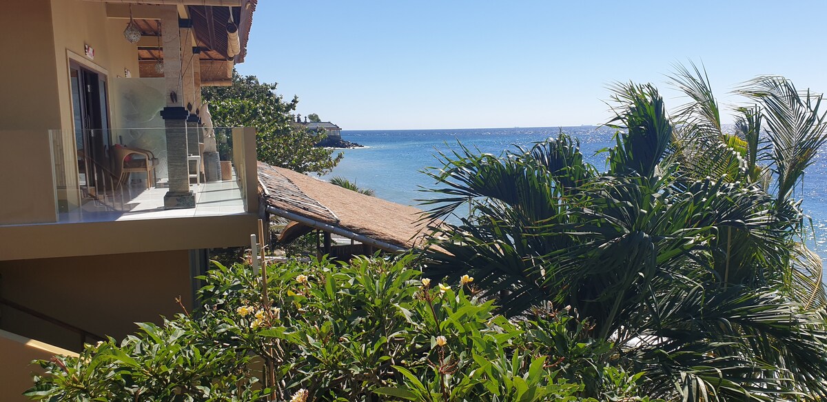 The image captures a view from the suite showcasing a lush greenery in the foreground, leading to the sparkling ocean in the background. A spacious balcony with seating is visible, alongside a charming thatched-roof structure to the side. Bright sunlight illuminates the scene.