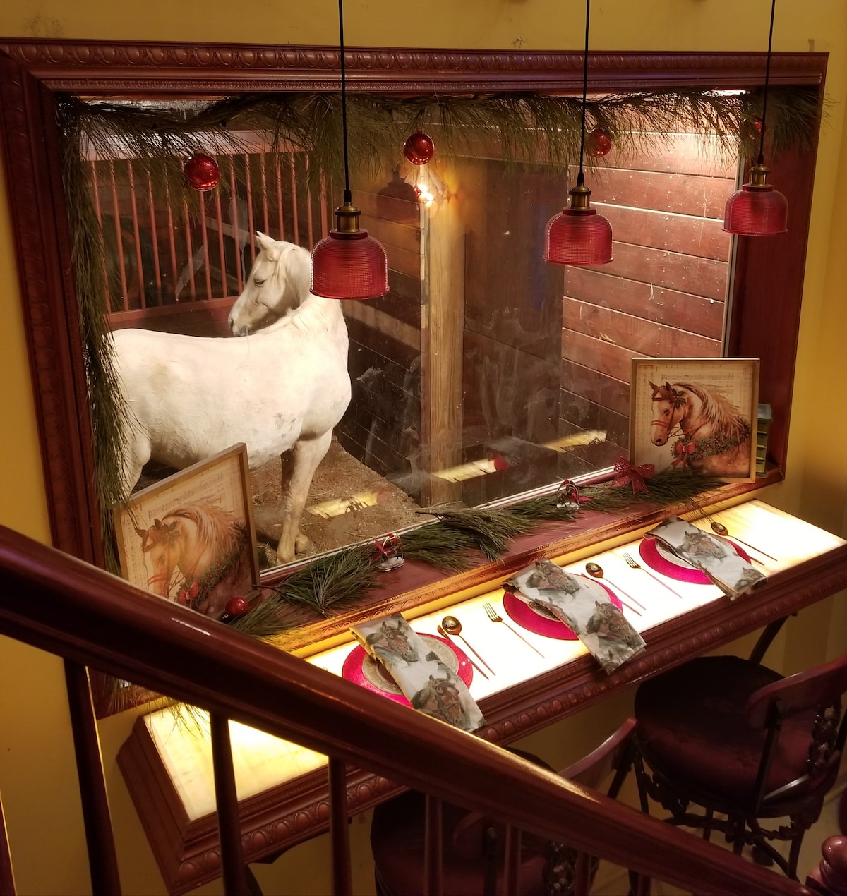 A decorative dining area is visible, featuring a polished wooden table set with plates and utensils. Above, three pendant lights with red shades provide illumination. A large window showcases a white horse in a stable, framed by festive greenery and artwork of horses.