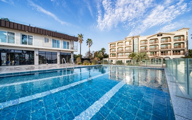 A large outdoor swimming pool is shown, featuring clear blue water reflecting the sky. Surrounding the pool are modern buildings with balconies and palm trees, contributing to a relaxed atmosphere. The expansive deck area provides ample space for lounging.