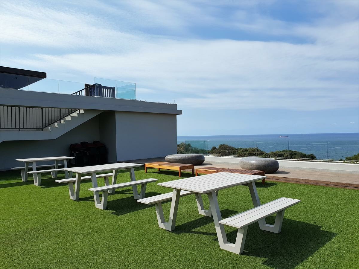 An outdoor communal area is displayed, featuring several picnic-style tables arranged on a green lawn. The ocean serves as a backdrop, with a clear blue sky and distant ships visible on the horizon. There are modern seating elements incorporated within the space.