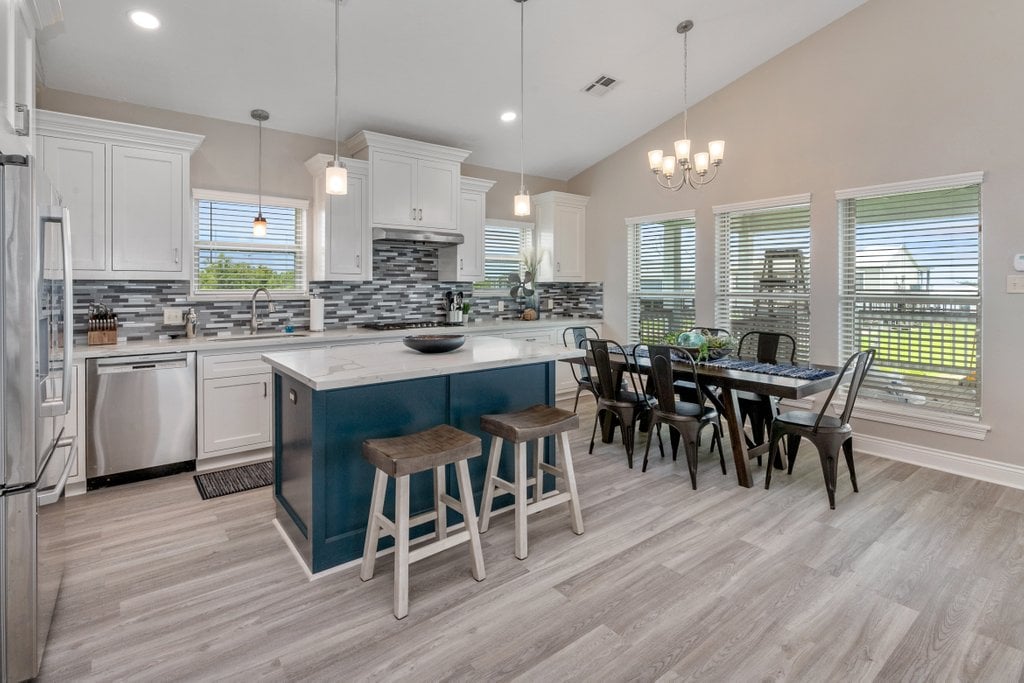 A modern kitchen and dining area features white cabinetry, a spacious island with wood stools, and a dining table surrounded by black chairs. Large windows allow natural light to flood the space, while a neutral color palette creates a clean and airy environment.