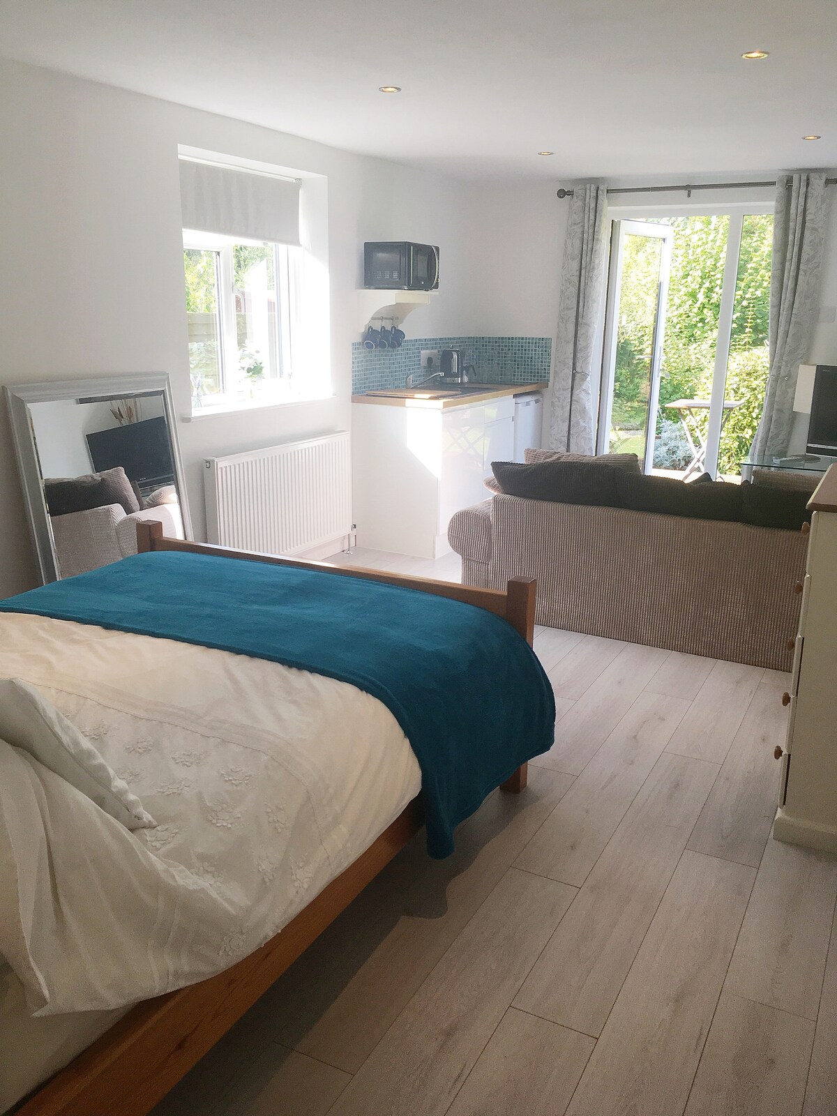 A bright studio space is furnished with a wooden bed draped in white linens and a teal throw. A cozy sofa is positioned beside a TV, with a small kitchenette featuring modern amenities. Natural light illuminates the area through large windows that overlook a garden.