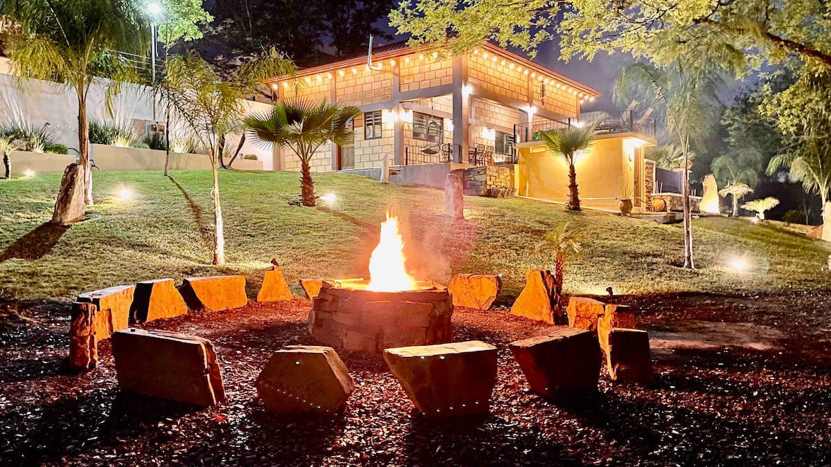 A fire pit surrounded by natural stone seating is centrally located in the green outdoor area. The warm glow of the fire contrasts with the illuminated exterior of the Cabaña, showcasing the inviting ambiance. Lush palm trees and landscaping enhance the serene environment.