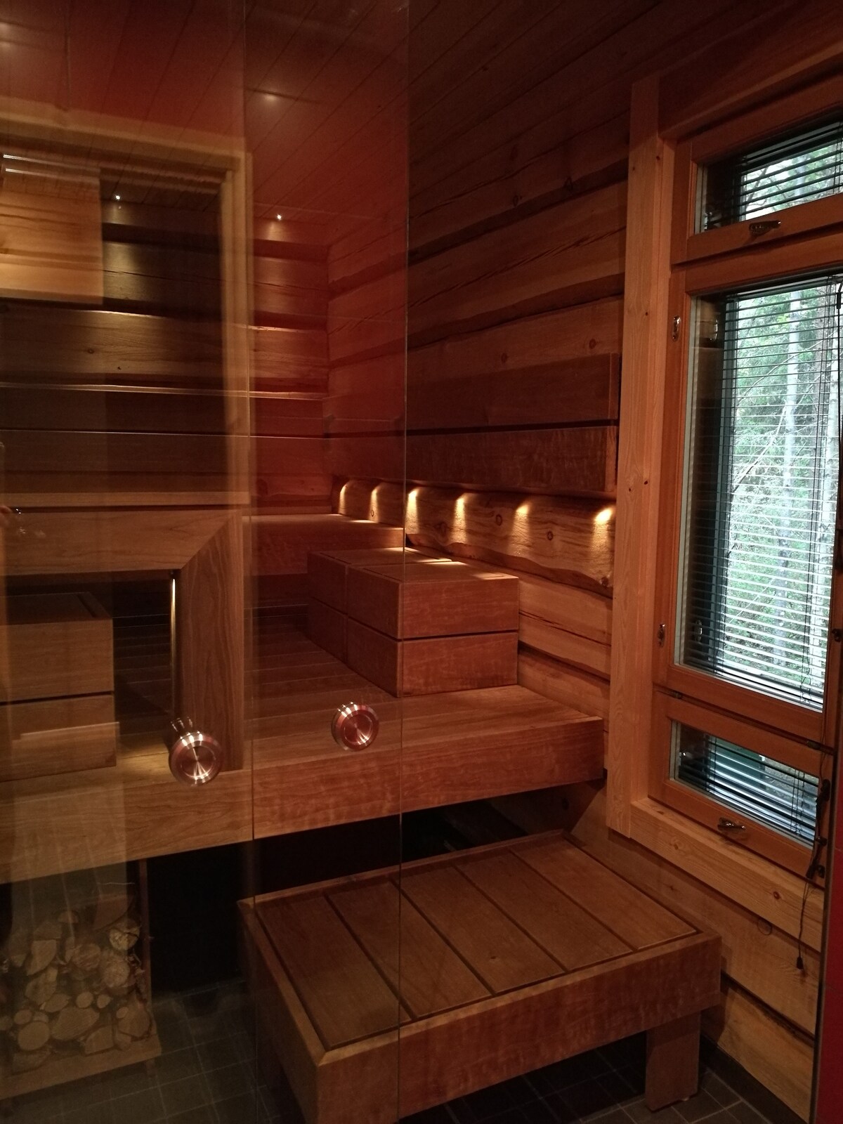 A wooden sauna is highlighted with warm lighting, showcasing smooth, natural wood paneling on the walls. Two wooden benches provide seating, with a sleek, wooden storage unit positioned in the center. Glass walls offer a view of the exterior, enhancing the open atmosphere.