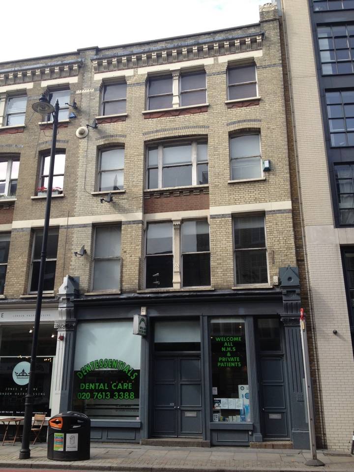 A three-story brick building features a mix of large windows and smaller panes, with a storefront at the ground level displaying signage. The facade incorporates light-colored bricks and dark framework, lending character to the structure. Urban amenities surround the building.