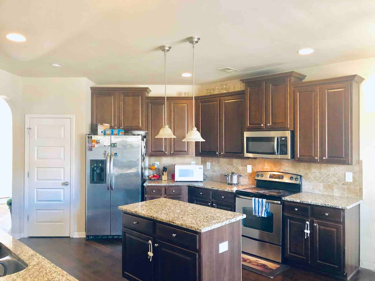 A modern kitchen is showcased with dark wooden cabinetry and granite countertops. Stainless steel appliances, including a refrigerator and oven, are visible. Pendant lights hang over a spacious central island, complementing the overall functionality of the space.