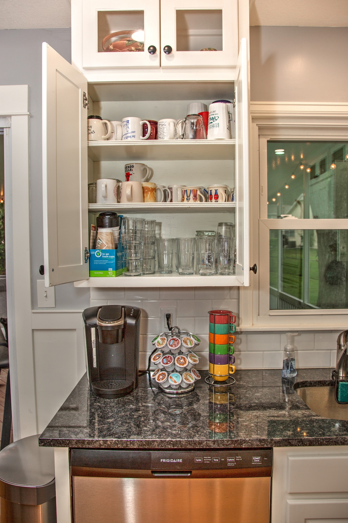 A well-organized kitchen area displays an array of glassware and mugs in a light-colored cabinet. A Keurig coffee maker is positioned on a dark granite countertop, with a carousel holding coffee pods nearby. Natural light filters through the window, enhancing the inviting space.