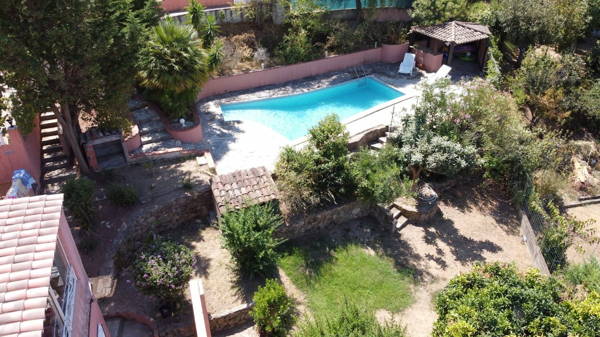 The image showcases an aerial view of a villa's outdoor space, featuring a swimming pool surrounded by lush greenery. A rustic gazebo is visible near the pool, along with comfortable lounge chairs. The landscape is characterized by well-maintained gardens and stone pathways.