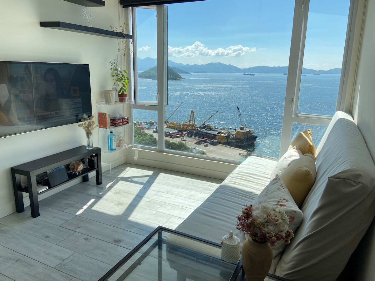 A bright living area is featured, showcasing a cozy sofa facing a large window with stunning views of the sea and distant mountains. Natural light illuminates the space, while a glass coffee table sits between decorative items, enhancing the inviting atmosphere.