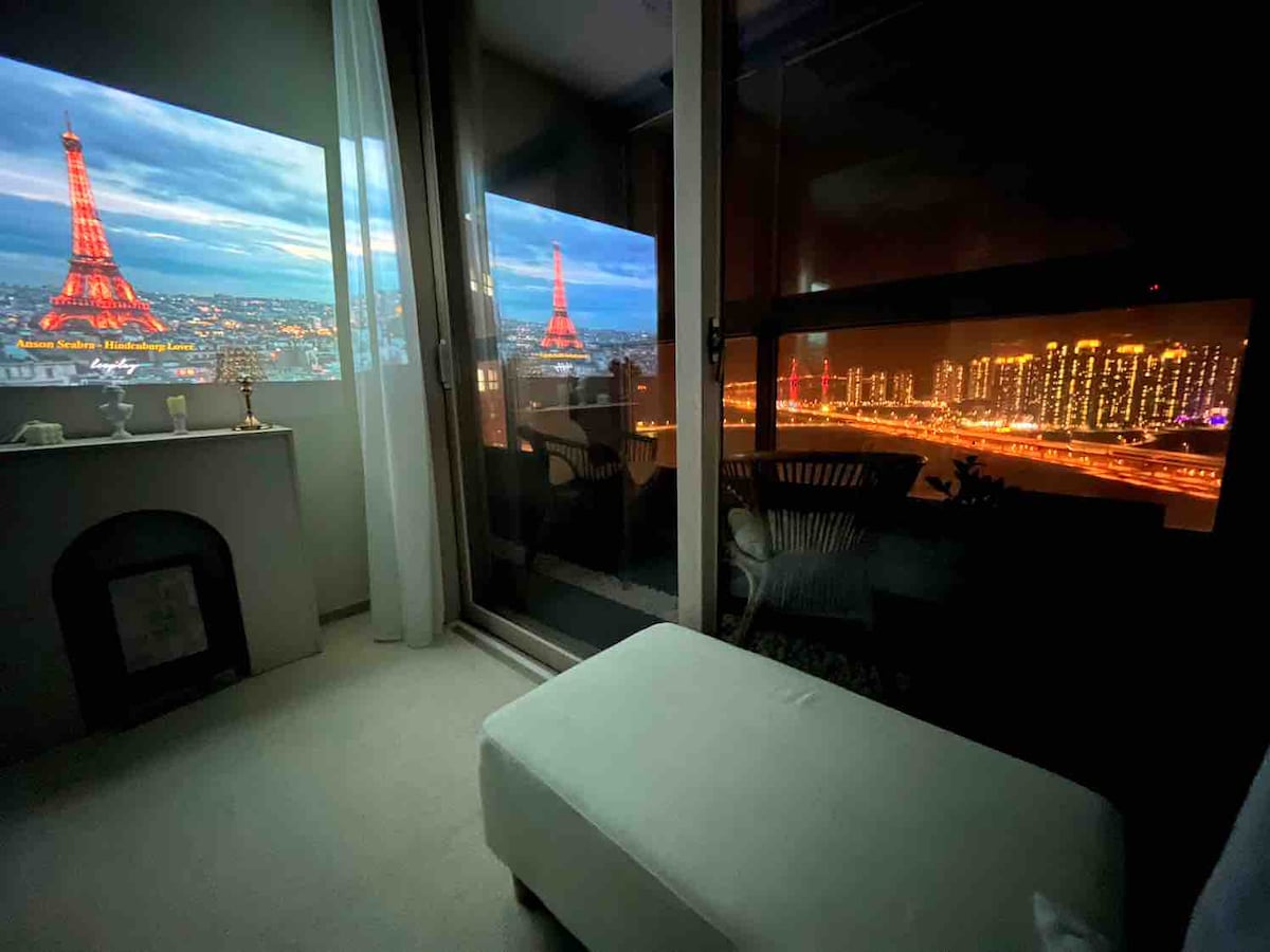 A cozy corner of a room features a light-colored seating area with a minimalist design. Large windows showcase stunning city views at night, featuring illuminated buildings and a prominent bridge. A projection of a landmark adds a unique touch to the ambiance.
