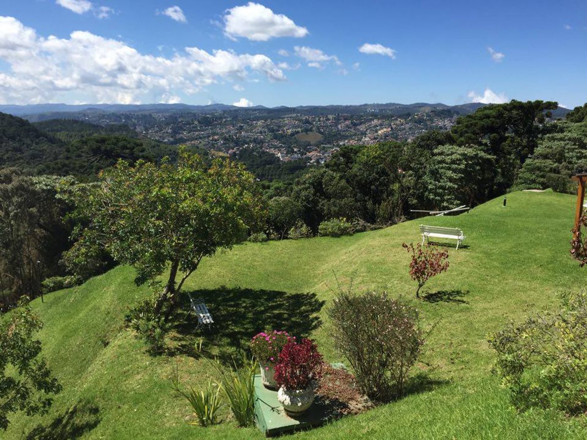 A spacious green lawn is set against a backdrop of rolling hills and a distant view of the town. Various trees and shrubs, including flowering plants, add natural beauty. A white bench is visible in the distance, inviting relaxation amidst the scenic landscape.