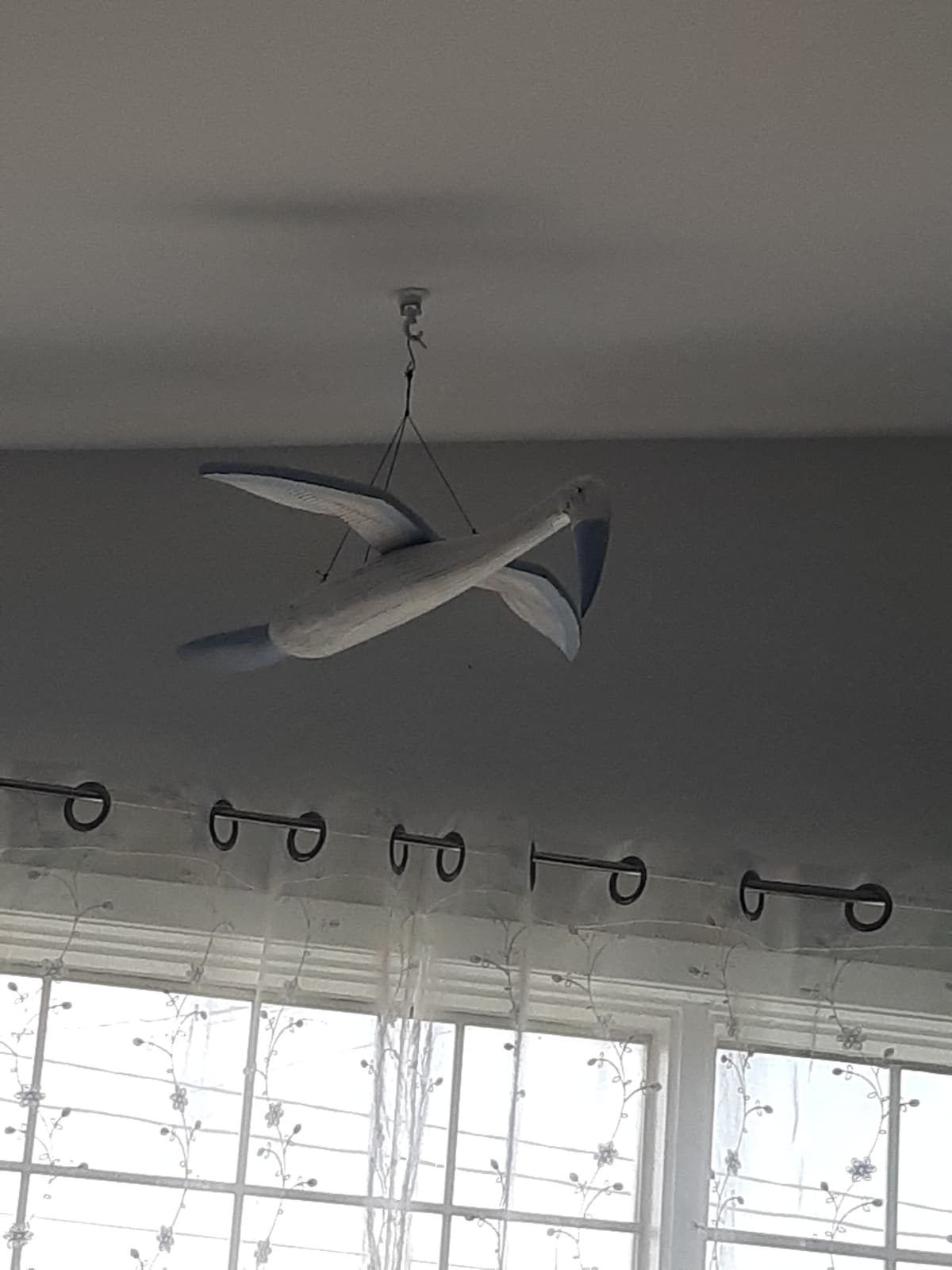 A decorative bird sculpture is suspended from the ceiling, showcasing a smooth, white finish with blue accents. Natural light enters through the nearby window, illuminating sheer curtains that softly frame the scene.