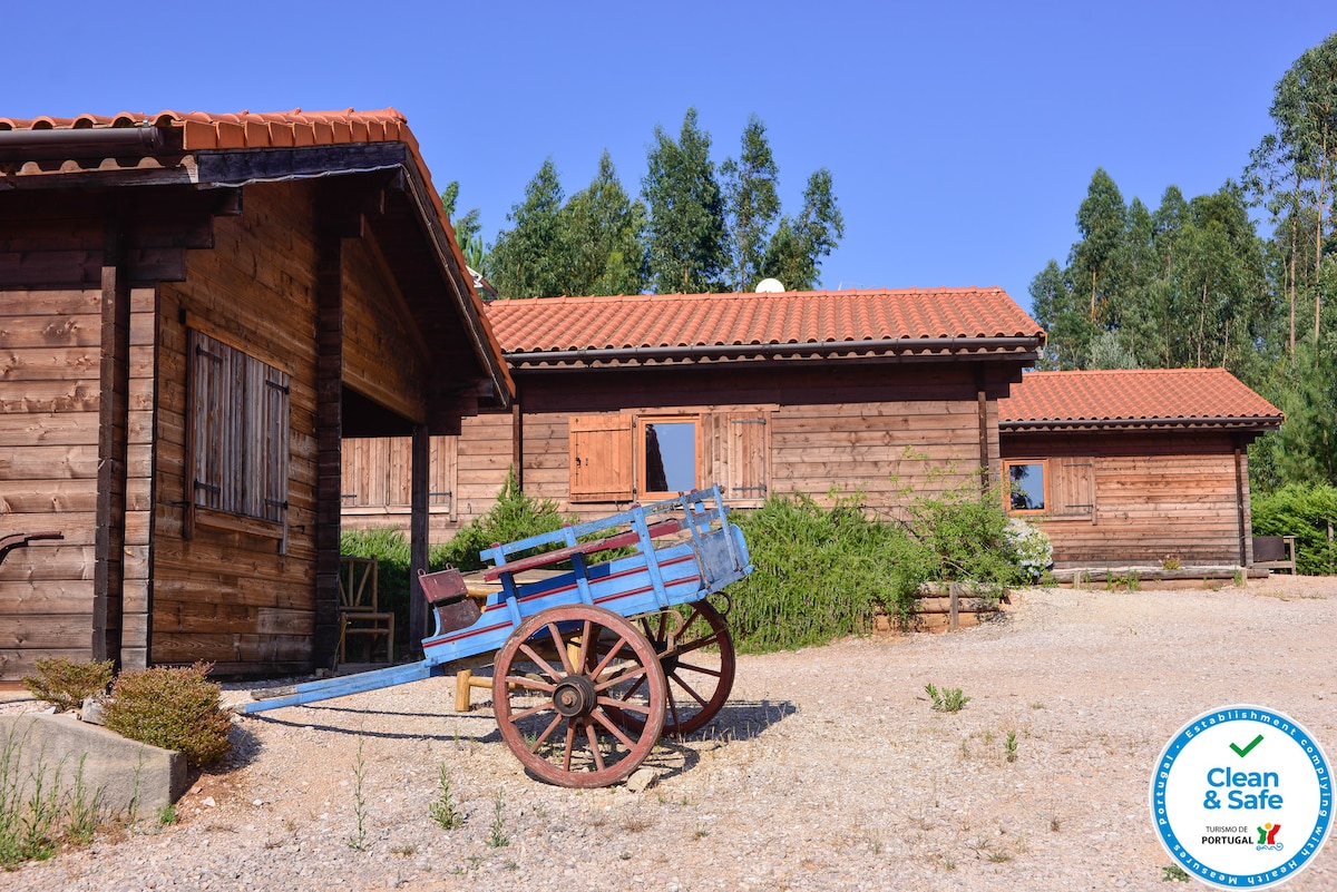 A rustic wooden house is positioned amidst lush greenery, featuring a traditional wooden cart in the foreground. The roof is tiled, and the exterior is complemented by shuttered windows, creating a serene and natural ambiance.