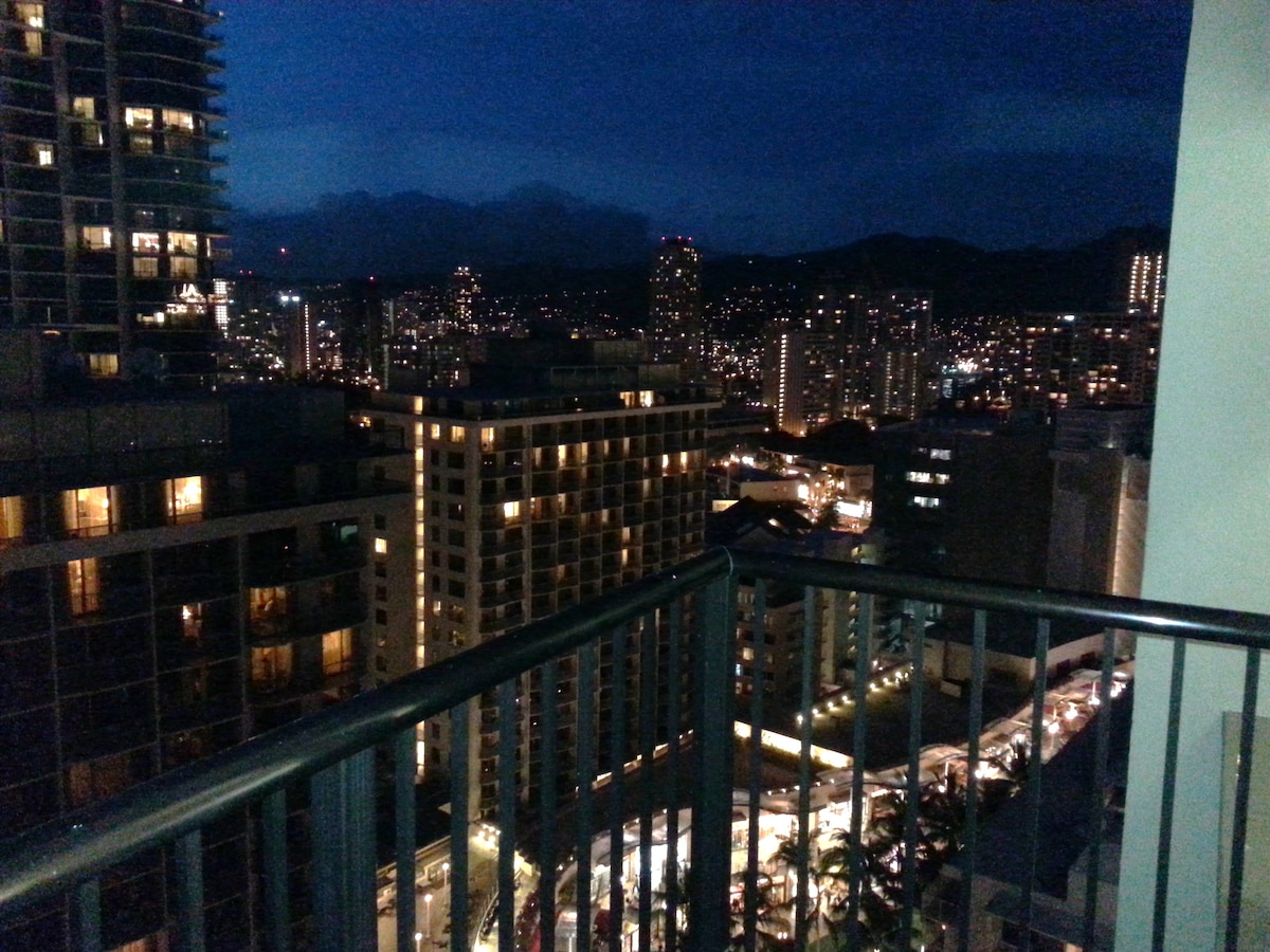 A cityscape view is captured at night, showcasing twinkling lights from buildings and streets below. The balcony railing frames the scene, while distant mountains are faintly visible against the night sky. This perspective highlights the vibrant atmosphere of Waikiki after dark.