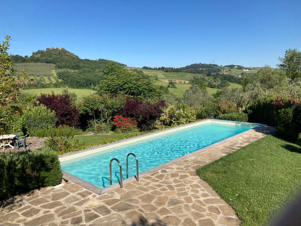 An exclusive swimming pool is surrounded by lush greenery and blooming flowers, offering serene views of the rolling hills in the distance. The poolside features stone flooring and steps leading into the clear water, inviting relaxation in a tranquil outdoor setting.