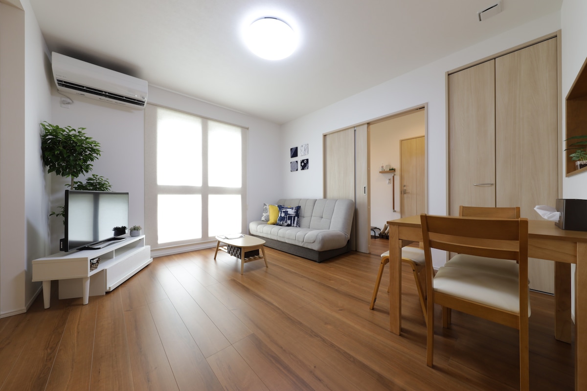 A well-lit living area features a comfortable sofa, accompanied by a lightweight coffee table. A television is positioned on a stylish unit, while natural light filters through large windows, illuminating the wooden flooring. A dining table set for four is positioned nearby.