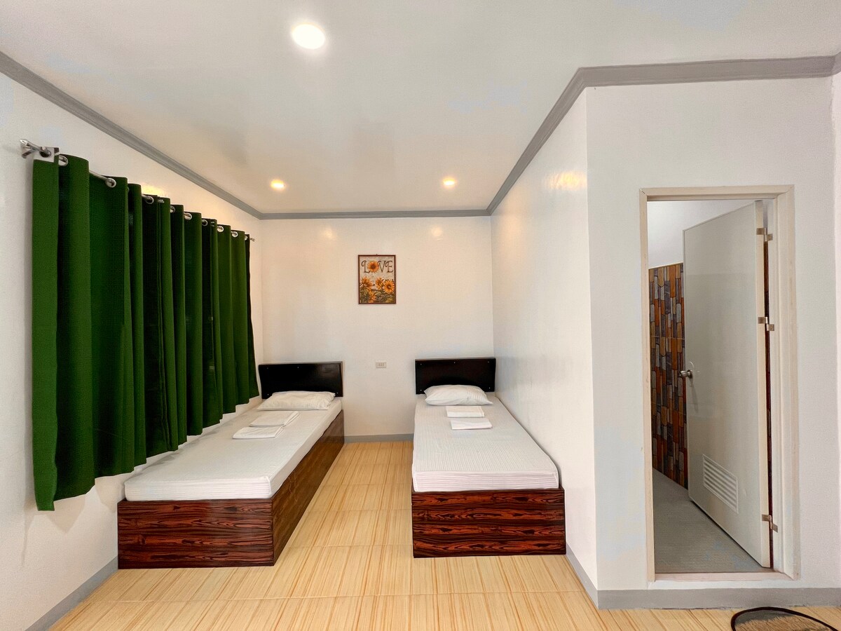 A simple bedroom features two twin beds with white linens arranged against a bright wall. Dark wood bed frames complement the neutral tones. Floor-to-ceiling green curtains frame a window, allowing natural light to brighten the space. A door leads to a bathroom.