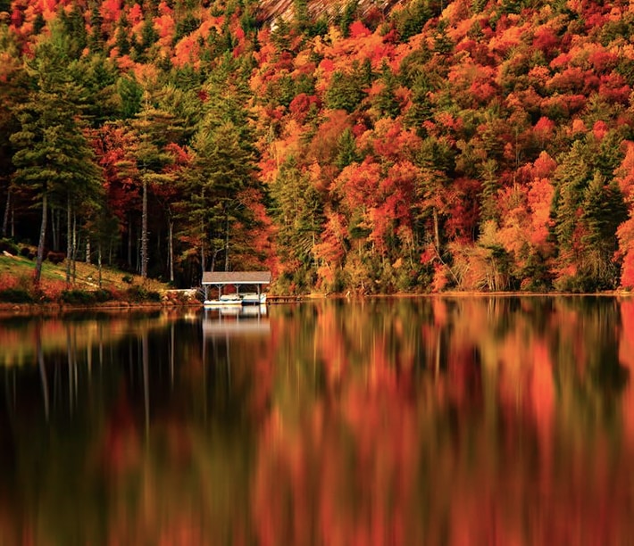 A serene lake reflects vibrant autumn colors, with trees displaying shades of red and gold surrounding the water. A small dock is visible, providing a tranquil spot for visitors to enjoy the natural beauty.