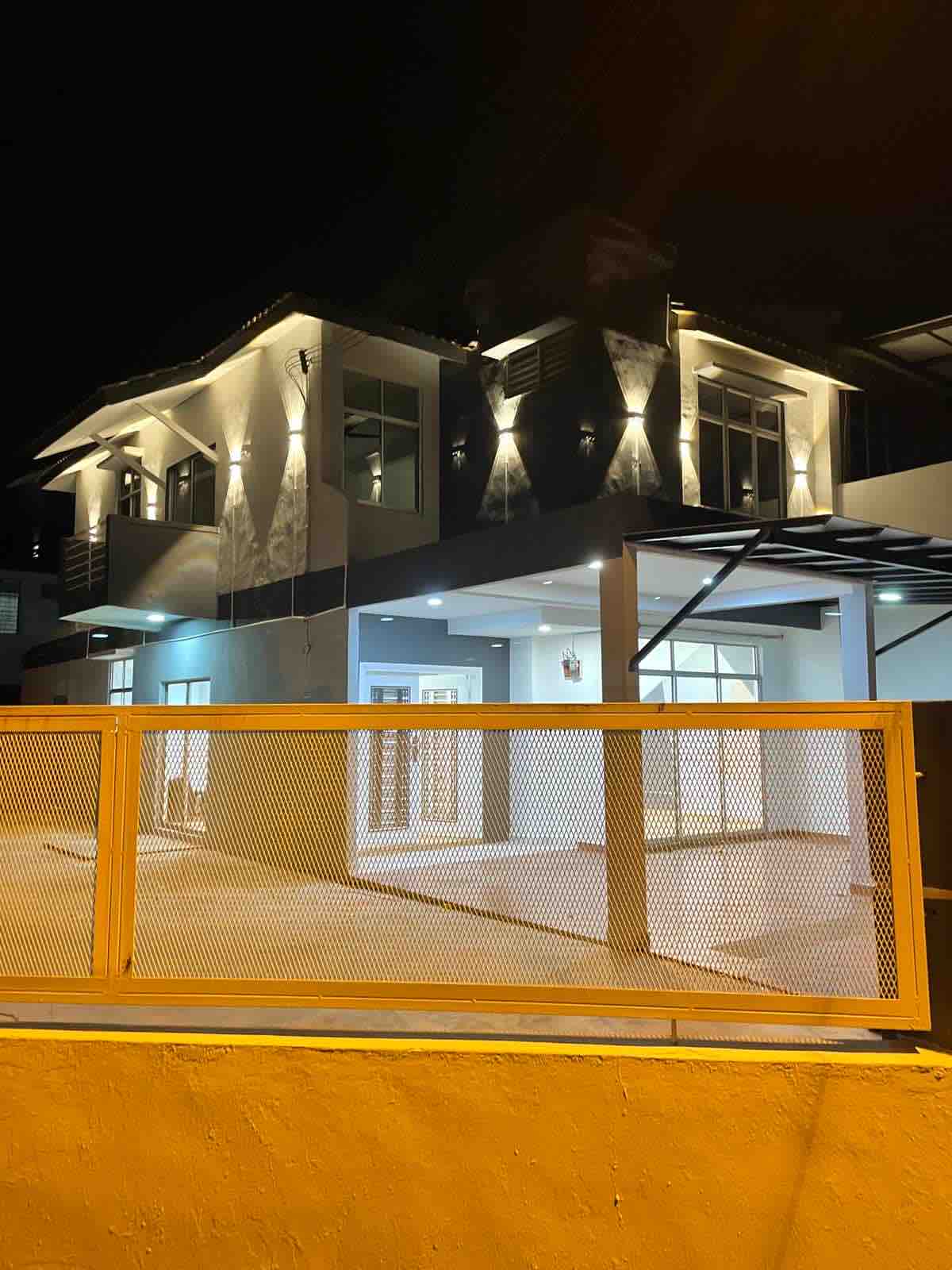 A modern two-story house is illuminated at night, displaying sleek architectural lines and a welcoming facade. The exterior features large windows and a covered area, while a stylish metal fence offers an overview of the property, enhancing its contemporary appeal.