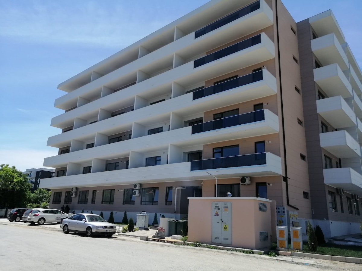 A newly constructed building with a modern facade is displayed. Multiple balconies extend from the upper floors, providing outdoor space. Large windows allow natural light to enter the living areas. The surrounding area includes a parking lot and landscaped greenery.