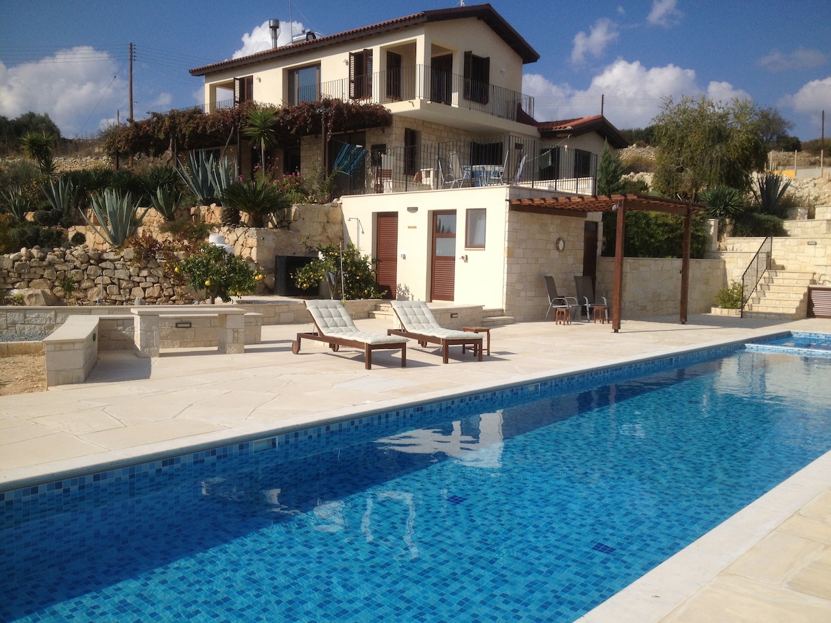 The image showcases a modern two-story villa surrounded by landscaped gardens. A large lap pool glistens in the foreground, with lounge chairs positioned nearby. The covered pergola and cabana room are seen adjacent to the pool, providing shaded areas for relaxation.