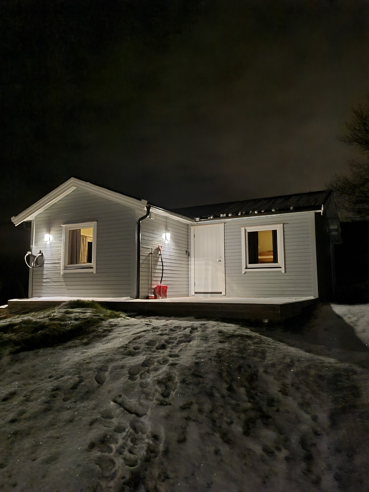 A charming, single-story house is illuminated at night, showcasing a cozy exterior with white wooden siding. Two windows are warmly lit, revealing the inviting interior. A small deck leads to the front door, while light snow covers the ground, reflecting the soft glow of the surrounding lights.