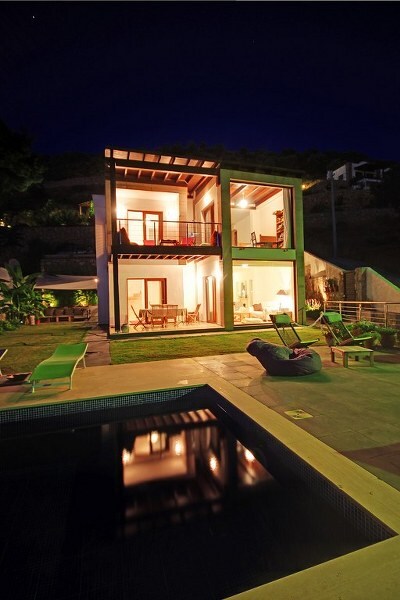 The villa is showcased at night, featuring a modern two-story design with large glass doors. Warm light spills from the windows, illuminating a spacious terrace with seating areas. A private pool reflects the surrounding scenery, creating a serene atmosphere in the garden setting.