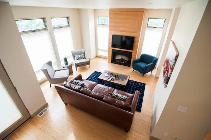The living area features a leather sofa facing a mounted television above an electric fireplace. Two armchairs in neutral tones are positioned beside large windows, allowing natural light to fill the space. A wooden coffee table, adorned with decorative items, completes the inviting atmosphere.