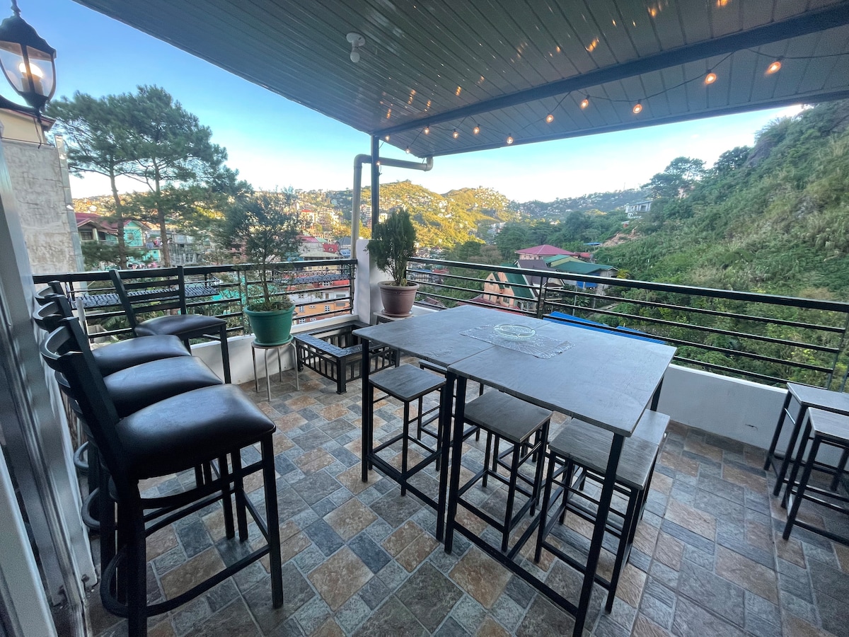 An outdoor terrace is furnished with a large table and multiple high-backed chairs, providing ample seating. Potted plants are positioned on the railing, while a scenic mountain view is visible in the background, creating a serene setting for relaxation.