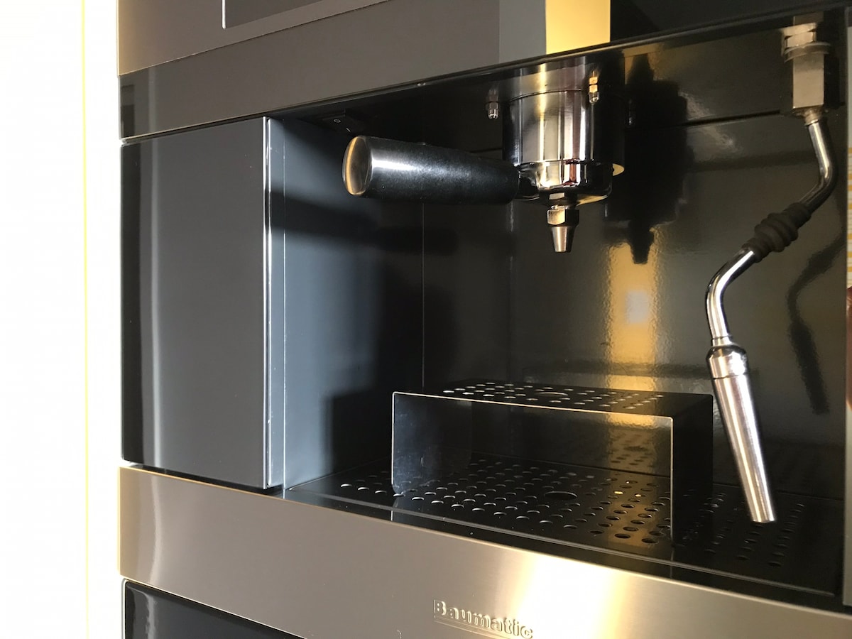 A built-in coffee machine is displayed with sleek black and stainless steel finishes. The front features a coffee spout and a metal steam wand, along with a drip tray positioned below. This appliance is designed for easy access and efficient use.