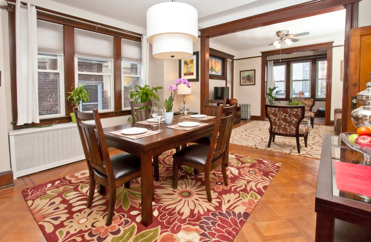 A dining area features a wooden table set for four, embellished with a floral tablecloth. Large windows allow abundant natural light, while potted plants enhance the space. In the background, a cozy living area with a patterned chair and decorative art adds a homey touch.