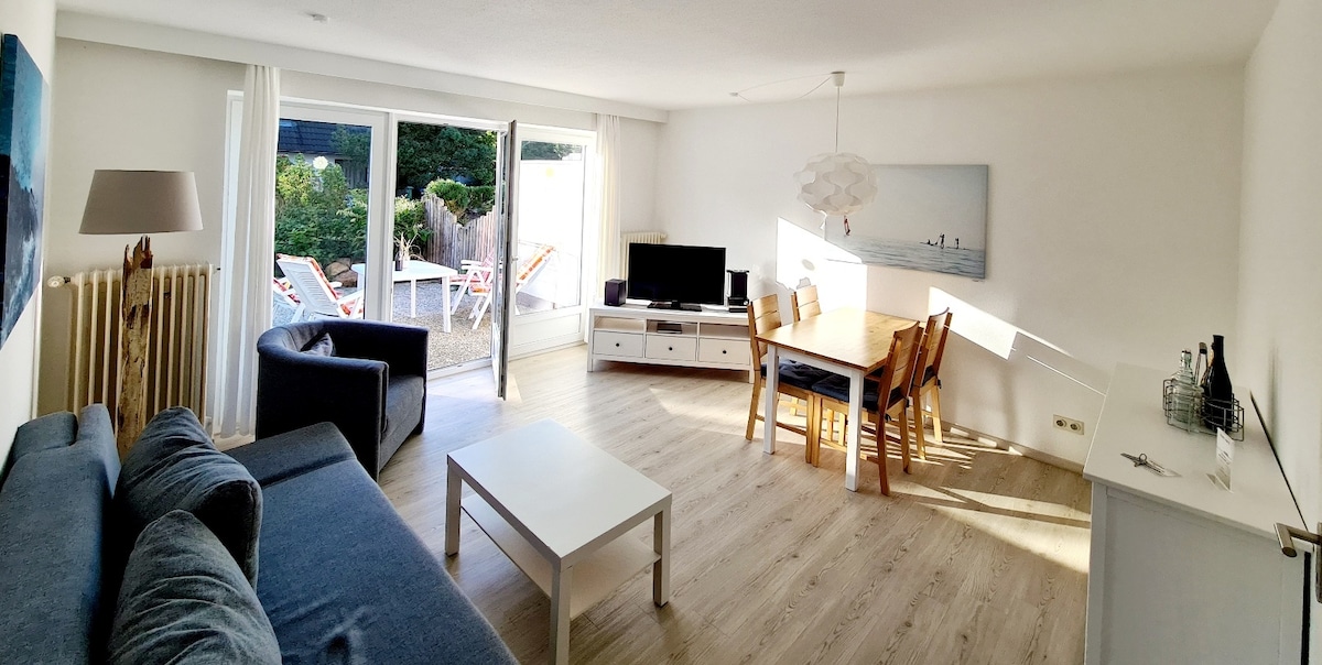 A bright and airy living space features a comfortable sofa and armchair, paired with a dining table for four. Large glass doors open to the terrace, allowing natural light to fill the room. A modern television is positioned on a white media console.