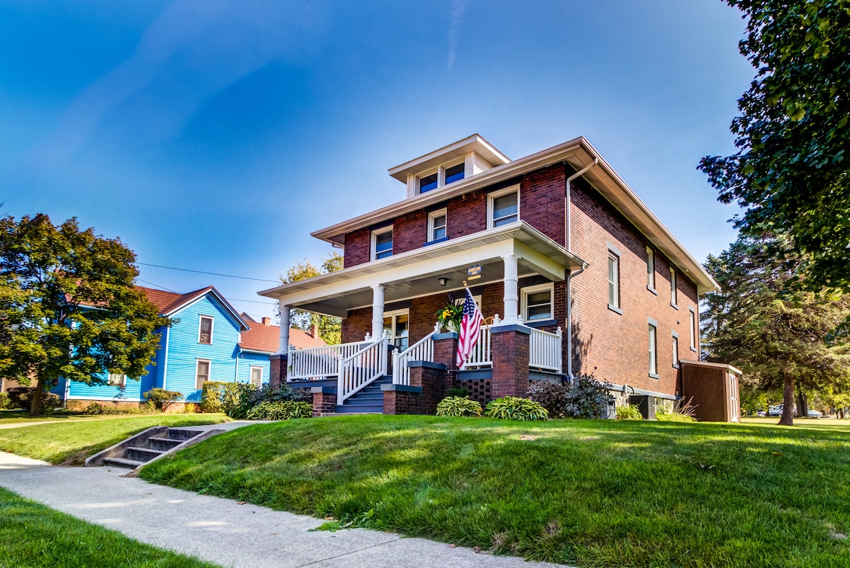 A charming historic home is showcased, featuring a welcoming front porch with white railings. Lush green grass surrounds the house, and colorful landscaping adds to the character. The two-story structure exhibits a classic brick façade, complemented by a vibrant blue house in the background.