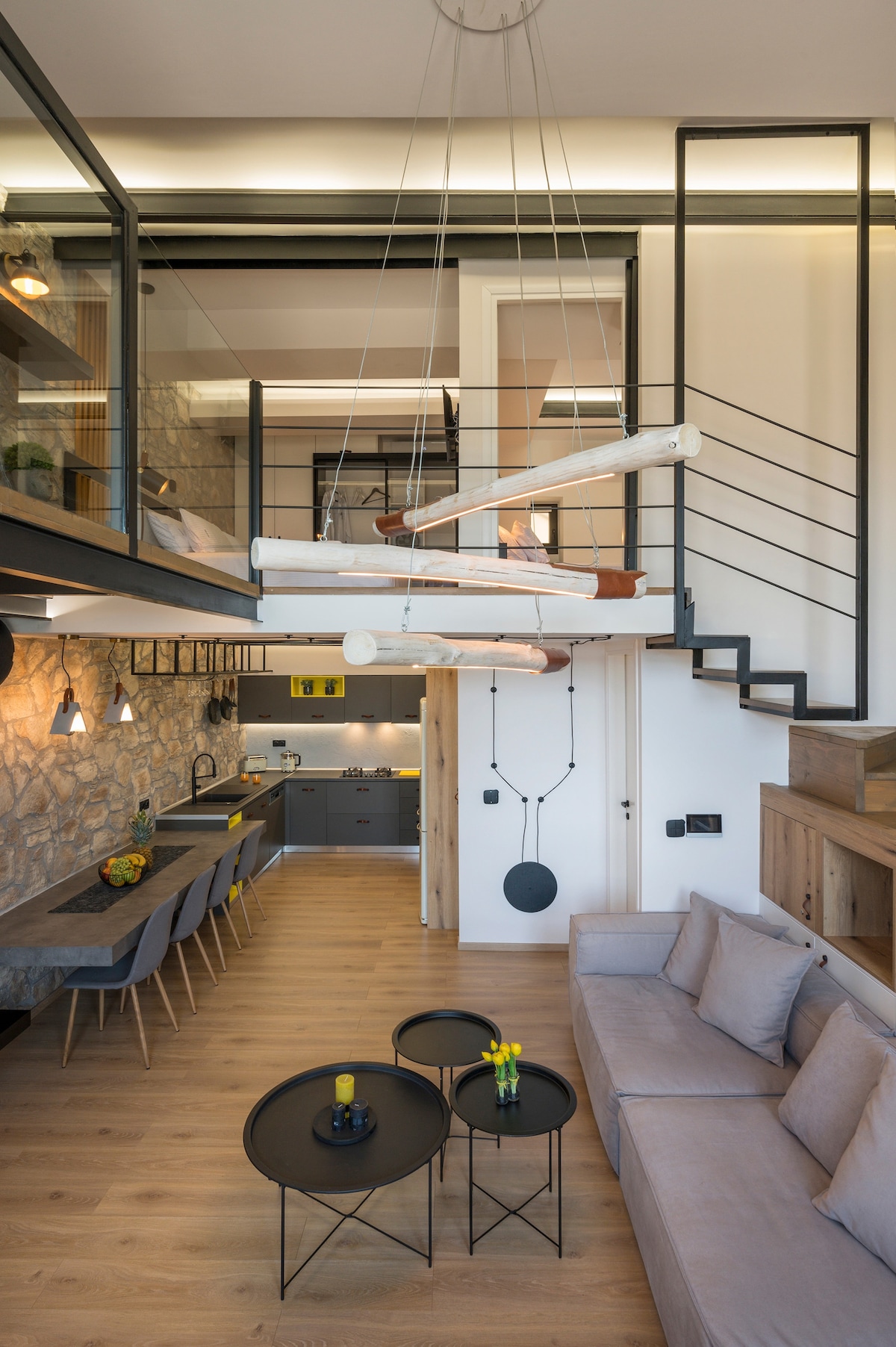 An open-concept living area features a modern design with warm wood tones and stone accents. A spacious sofa is paired with two circular coffee tables, while a dining area is equipped with a long table and several chairs. A staircase leads to a lofted space above.