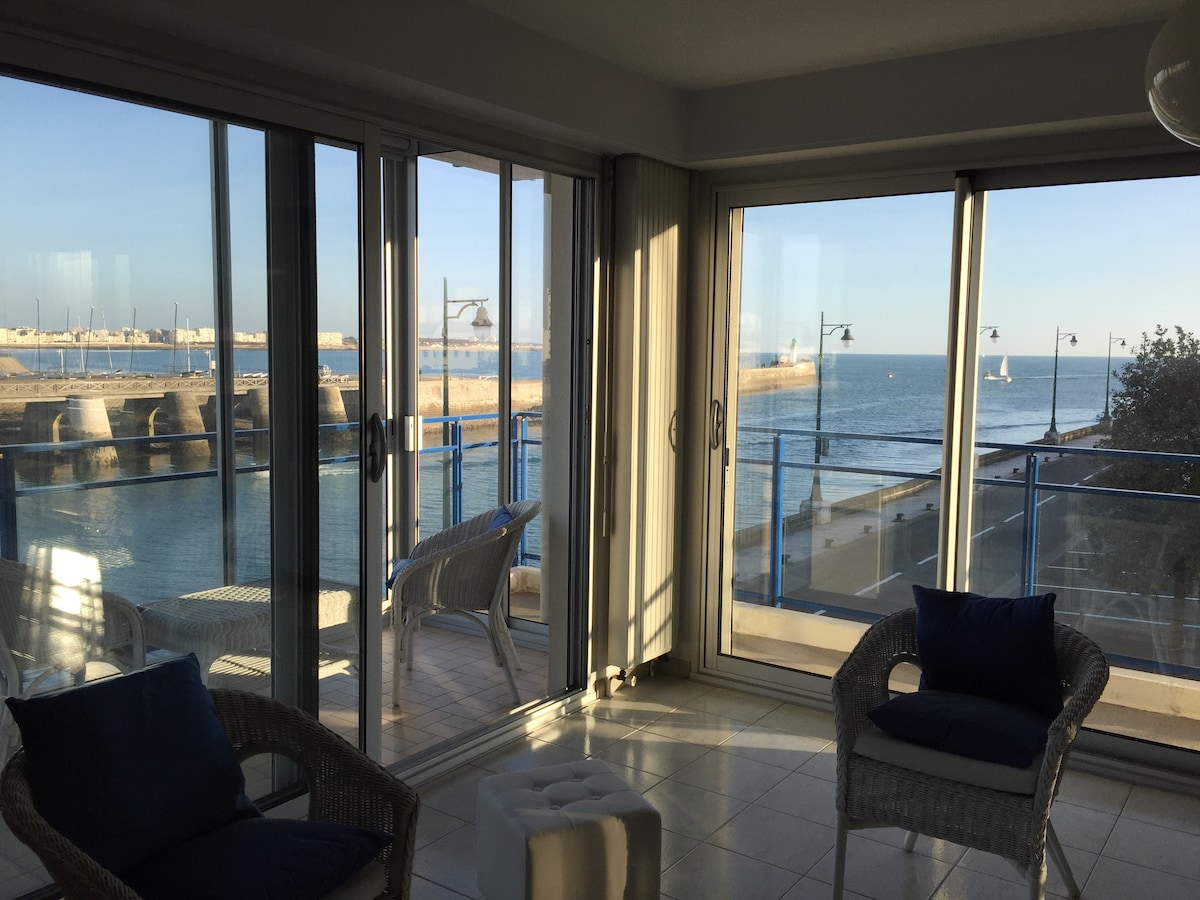 A light-filled living area features large glass doors that open to a view of the bay. Two wicker chairs are positioned beside a small ottoman, inviting relaxation while enjoying the scenic waterfront. The soft colors and neutral decor create a peaceful ambience.