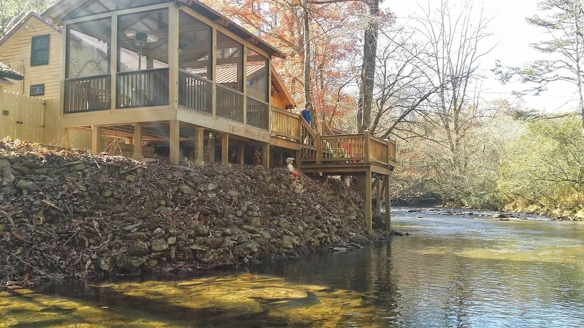 A cabin is positioned on the banks of a gently flowing creek, with a wooden deck extending over the water. The screened porch is visible, surrounded by trees displaying autumn foliage, creating a serene and natural setting.