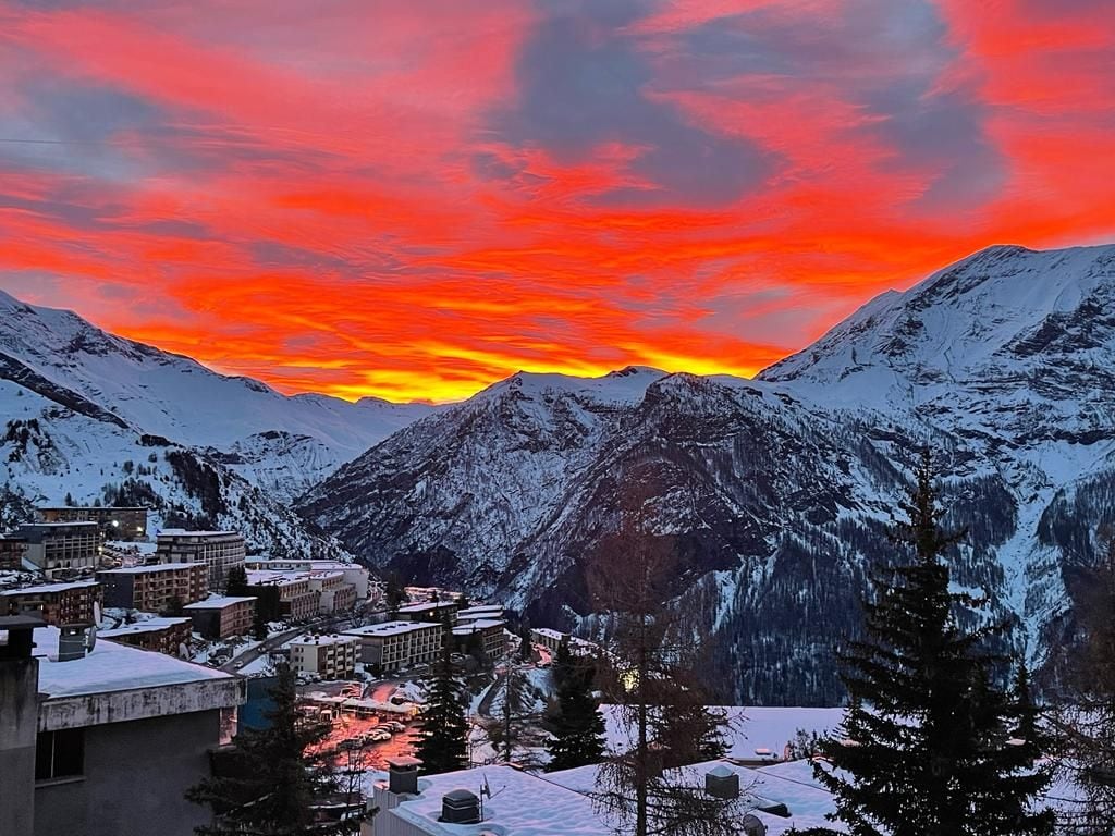 A breathtaking sunset paints the sky in vibrant shades of orange and pink, contrasting against the snowy mountains and valley below. The scene includes a view of the nearby village and trees, creating a serene winter landscape.