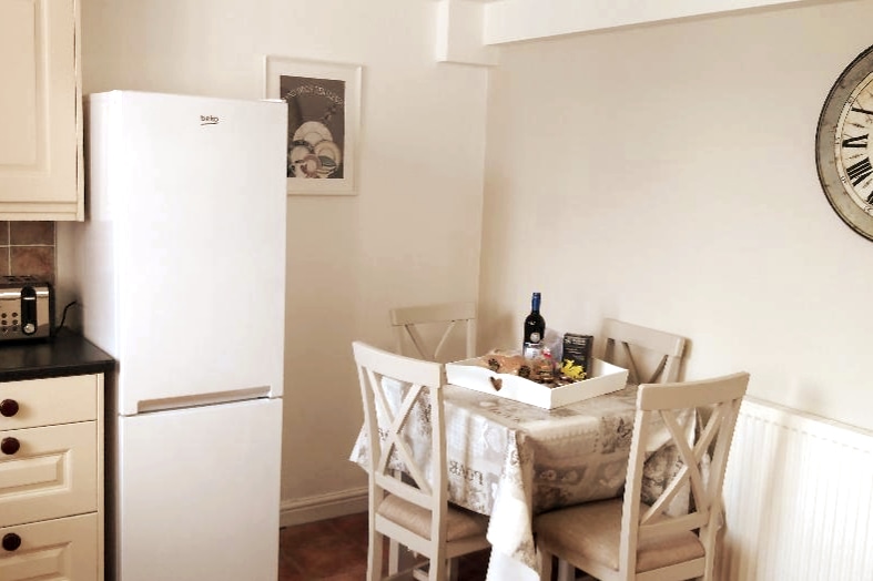 The kitchen area includes a white refrigerator and a small dining table set for four. A decorative clock and framed artwork adorn the walls, while a tablecloth adds a touch of warmth to the space.