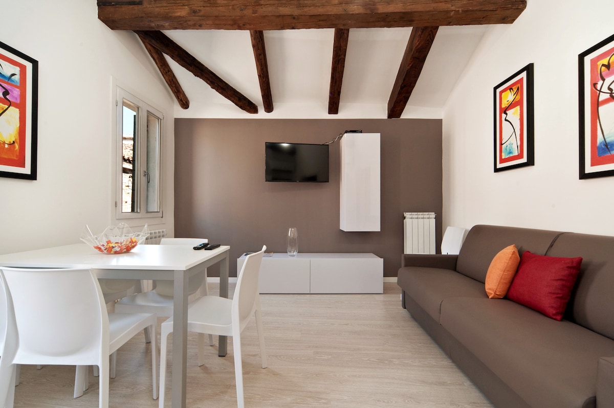 A modern living area features a spacious, muted brown sofa with a decorative pillow in red. Across from it, a white cabinet hosts a flat-screen television. A dining table with four white chairs is set nearby, complemented by framed artwork on the walls.
