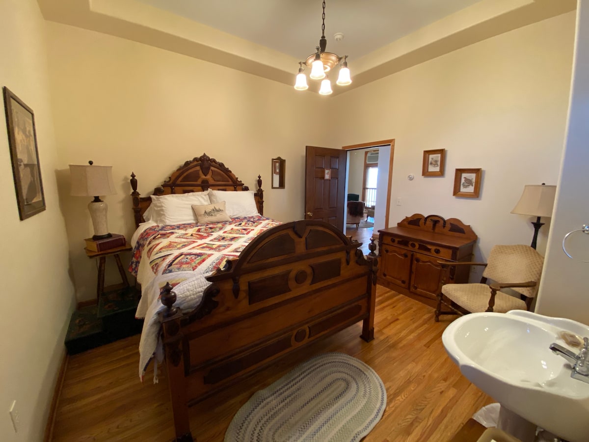 A historic bedroom features an ornate wooden bed with a quilted coverlet, complemented by two bedside lamps. A vintage dresser and a chair are positioned near the door. A cozy area rug lies on the hardwood floor, and natural light fills the room.