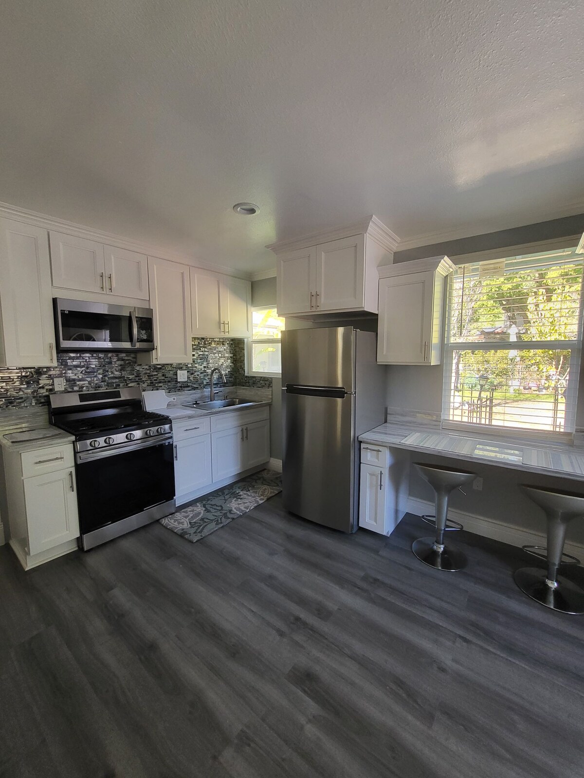 The kitchen area features modern cabinetry in white with a complementary backsplash. Stainless steel appliances, including a refrigerator and a gas stove, are present. A small dining space with two bar stools is positioned near a window that allows for natural light.