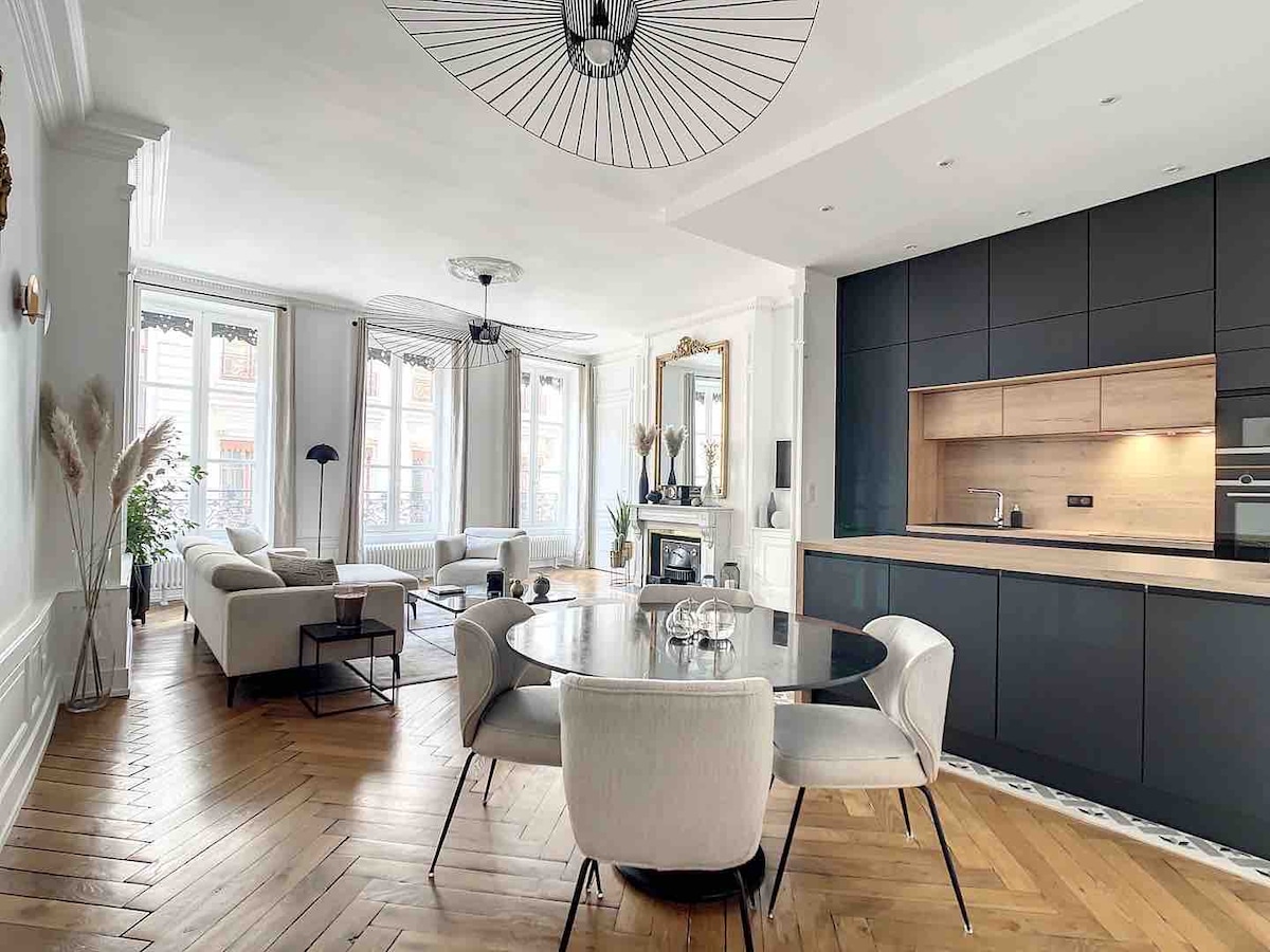 An open-plan living space features a dark kitchen area and a circular dining table set for four. A comfortable seating arrangement with plush sofas creates a welcoming atmosphere. Large windows allow natural light to fill the room, highlighting the polished wooden flooring.