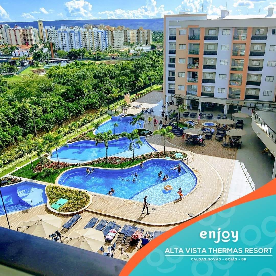 A view of the resort's pool area is presented, showcasing multiple swimming pools surrounded by lush greenery. Lounge chairs are arranged around the pools, with guests enjoying the water. The backdrop features a cluster of apartment buildings, illustrating the resort's spacious environment.