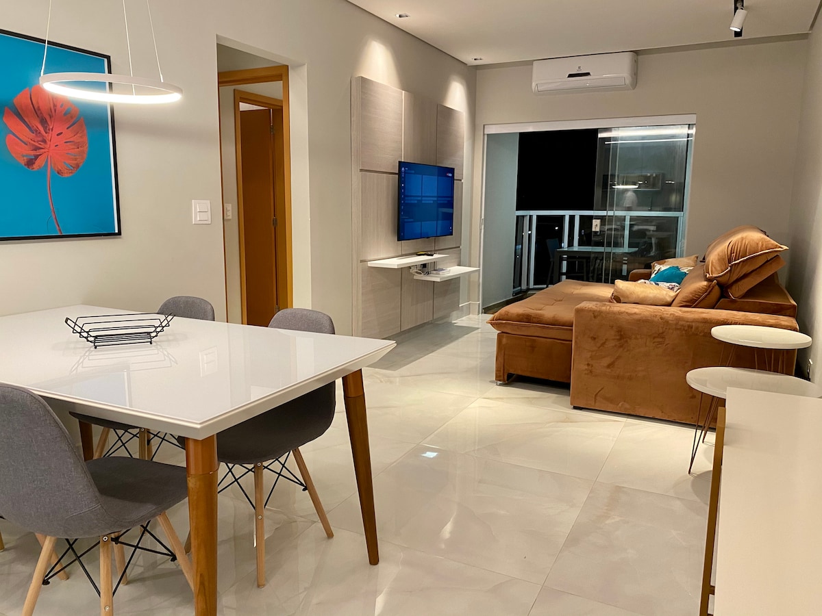 An open living and dining area features a clean, modern aesthetic. A white dining table with gray chairs sits beside a spacious sofa. A wall-mounted television is visible, and sliding glass doors lead to a balcony, providing natural light to the space.