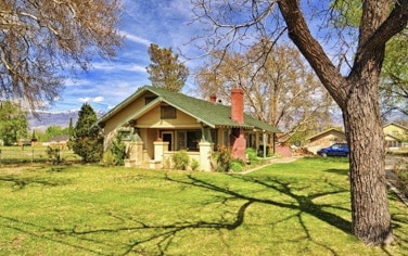 The Craftsman style house features a green roof and a red brick chimney, surrounded by lush greenery. Large trees provide shade, and the expansive lawn enhances the outdoor space. The building's charming architecture is complemented by a welcoming front porch.
