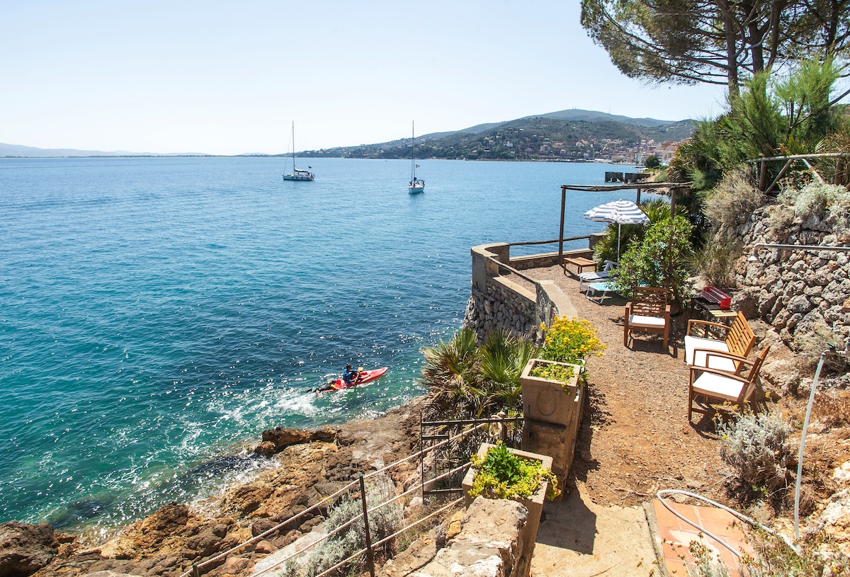 A serene outdoor space features rocky beach access with a kayak floating nearby. Comfortable lounge chairs are positioned on the terrace overlooking the sea, surrounded by coastal vegetation. The clear blue water reflects the sunlight, enhancing the tranquil atmosphere of the seaside setting.