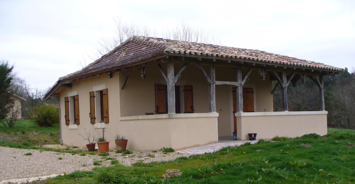 A simple one-story house is featured, built with a rustic design. The exterior consists of light-colored walls and a sloped roof made of tiles. Wooden shutters are placed on multiple windows, and a covered porch area is present, supported by wooden beams. Surrounding greenery adds a natural setting.